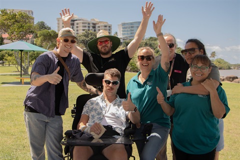 A group of seven people from the Brain Injury Community smiling and giving a 'thumbs up' gesture in a park at Jack Evans Boat Harbour, Tweed Heads. One member, seated in a wheelchair, joins in with a 'thumbs up,' surrounded by six others standing with their arms raised or also signalling 'thumbs up