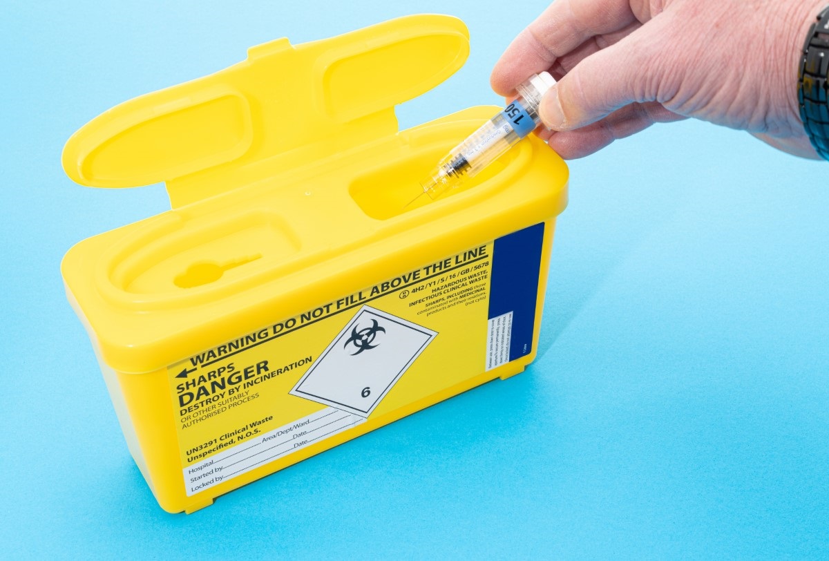 Hand putting used syringe into yellow sharps box.